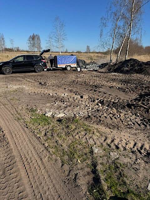 Czarny samoch&oacute;d typu SUV z&nbsp;przyczepką zaparkowany na&nbsp;piaszczystym terenie w&nbsp;okolicach Mrzeżyna. Na ziemi obok przyczepki widoczna jest sterta wyrzuconego gruzu budowlanego i&nbsp;odłamk&oacute;w betonu. W tle rosną brzozy pod bezchmurnym, błękitnym niebem.